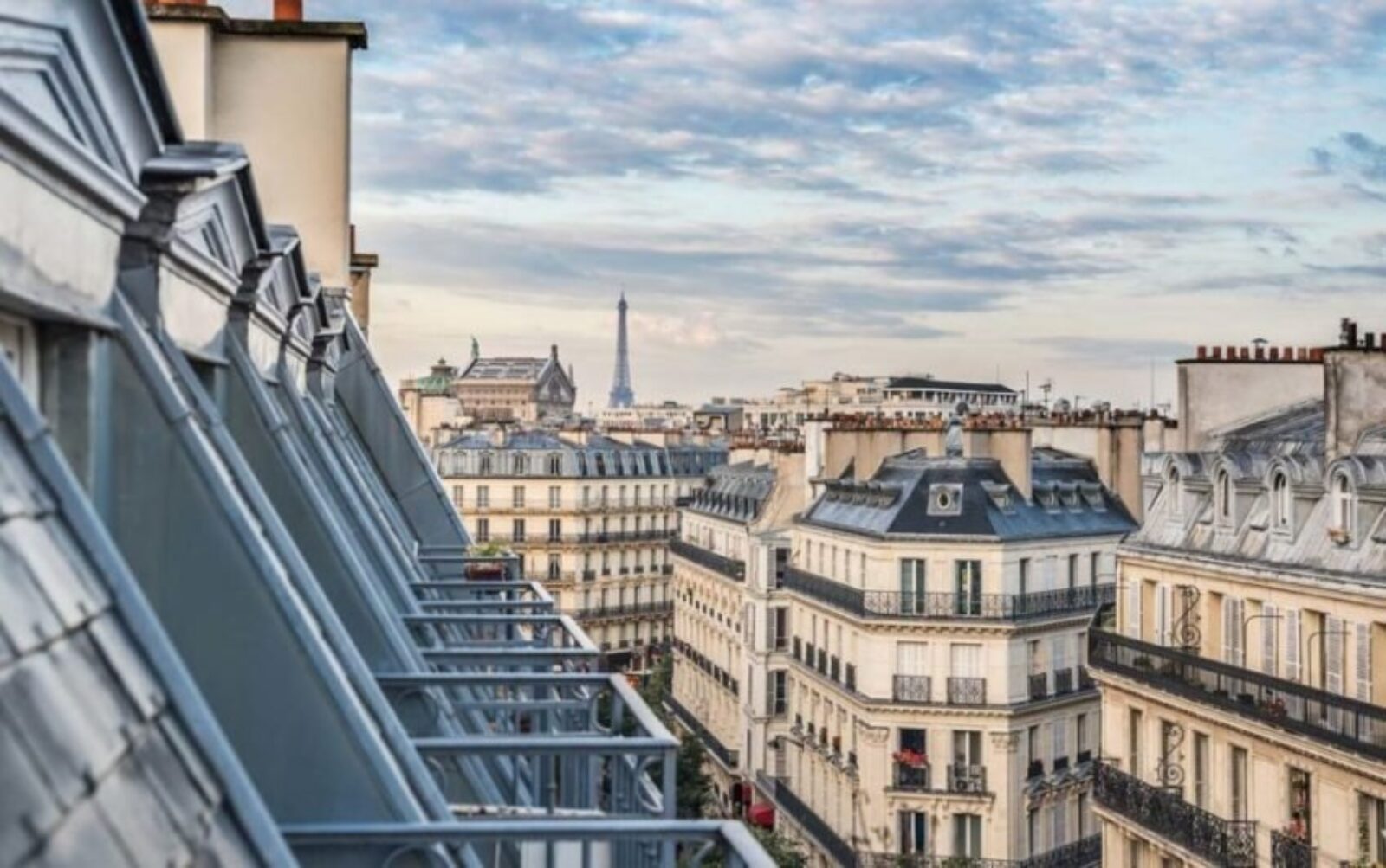 Paysage urbain parisien avec immeubles classiques de style haussmannien dotés de toits mansardés et de lucarnes. La Tour Eiffel s'élève au loin sous un ciel partiellement nuageux, créant une silhouette typiquement parisienne.