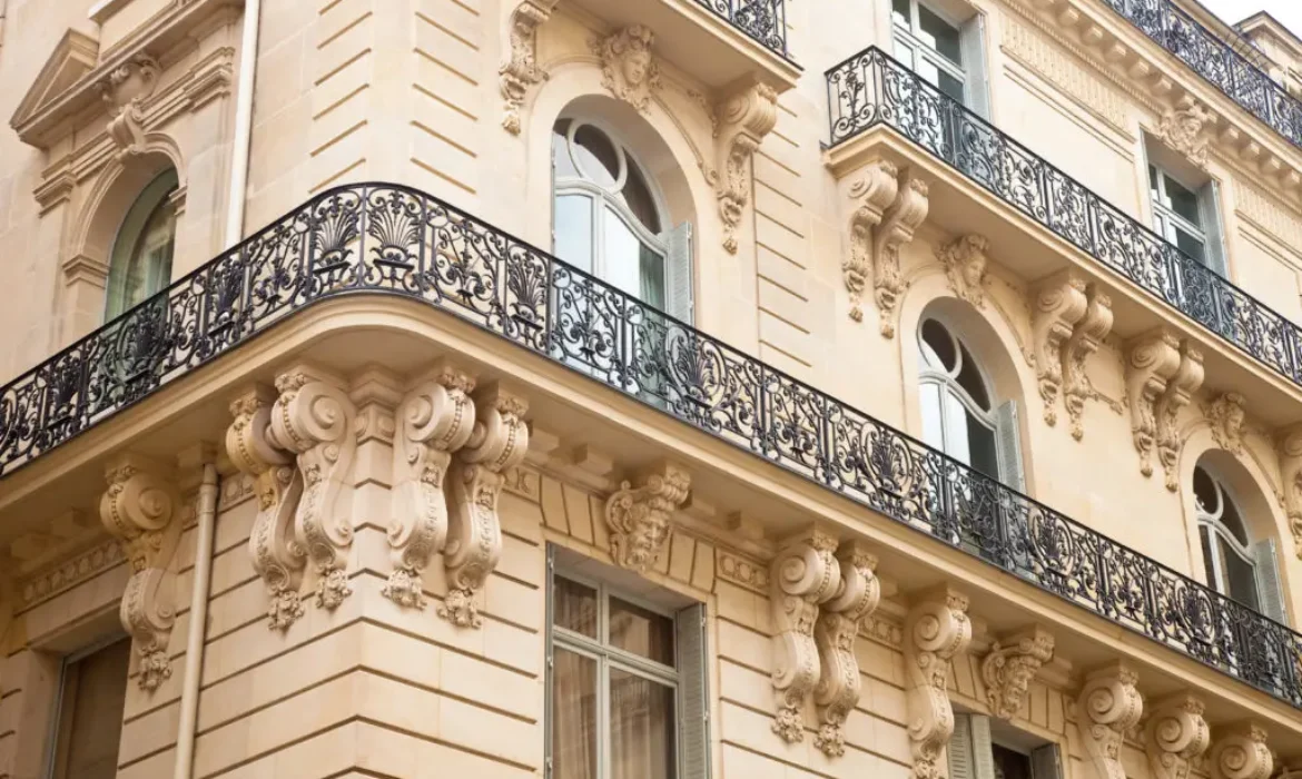 Vue en angle d'un bâtiment beige et orné de style européen. Il comporte plusieurs étages avec des balcons décoratifs en fer forgé, de grandes fenêtres cintrées et des détails sculpturaux complexes sur la façade, mettant en valeur une conception architecturale élégante.