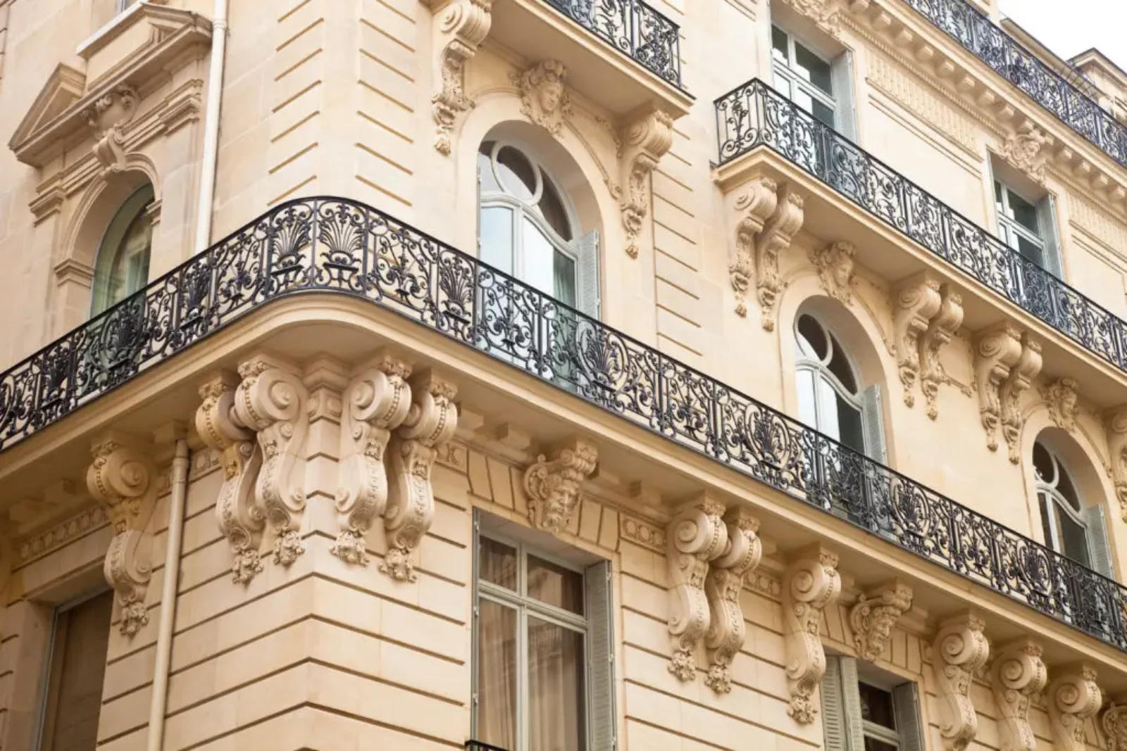 Vue en angle d'un bâtiment beige et orné de style européen. Il comporte plusieurs étages avec des balcons décoratifs en fer forgé, de grandes fenêtres cintrées et des détails sculpturaux complexes sur la façade, mettant en valeur une conception architecturale élégante.