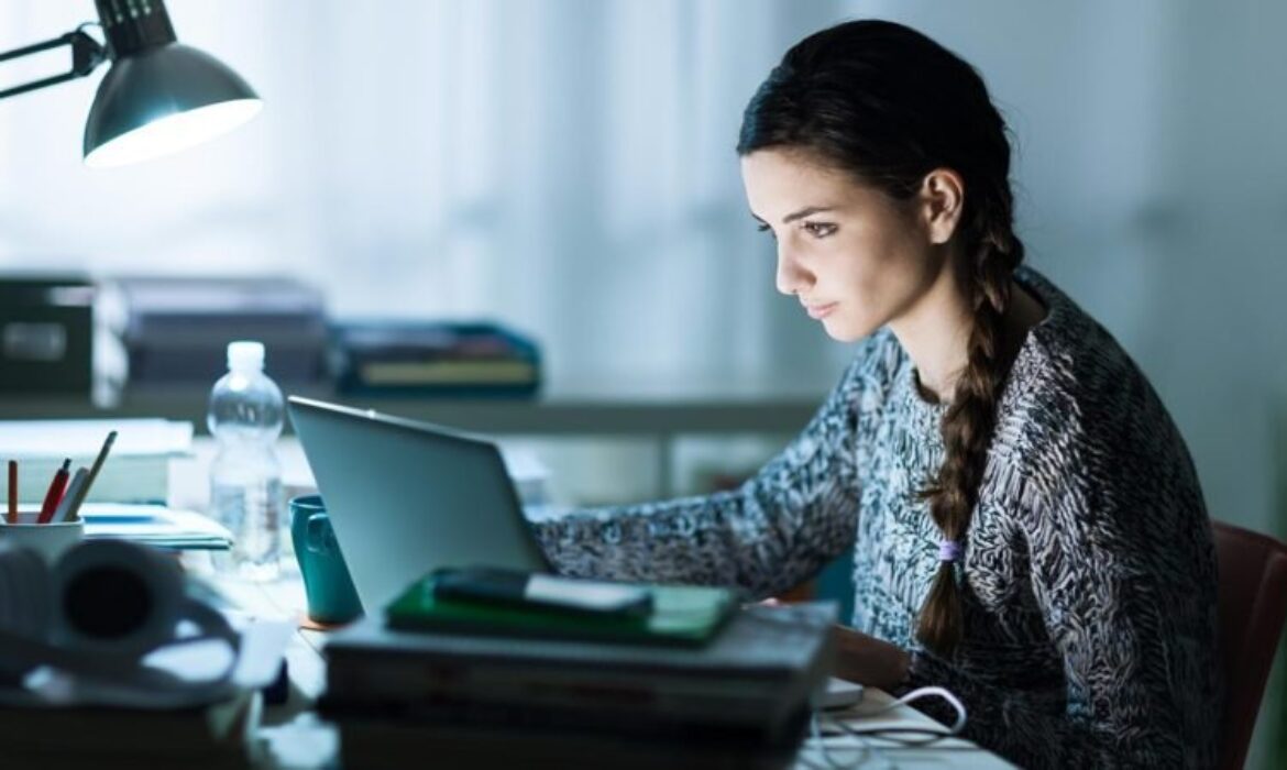 Une femme avec une longue tresse est intensément concentrée sur l'écran de son ordinateur portable alors qu'elle est assise à un bureau encombré de livres et de papiers, cherchant comment anticiper son achat immobilier à la lumière du coronavirus. La pièce est faiblement éclairée par une lampe de bureau. Une bouteille d’eau et quelques objets épars se trouvent également sur le bureau.