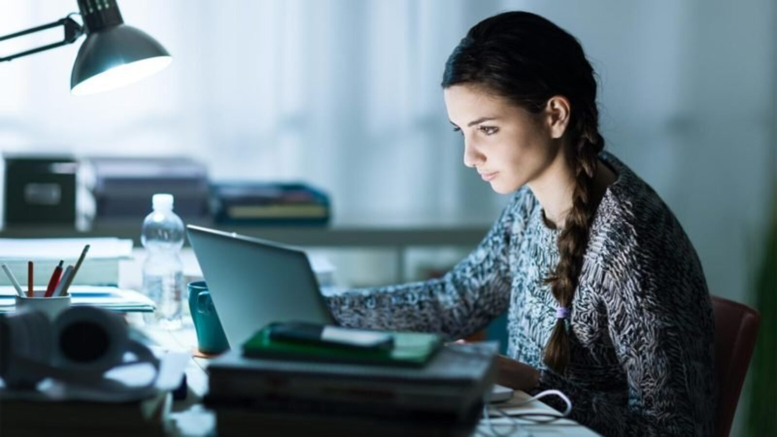 Une femme avec une longue tresse est intensément concentrée sur l'écran de son ordinateur portable alors qu'elle est assise à un bureau encombré de livres et de papiers, cherchant comment anticiper son achat immobilier à la lumière du coronavirus. La pièce est faiblement éclairée par une lampe de bureau. Une bouteille d’eau et quelques objets épars se trouvent également sur le bureau.