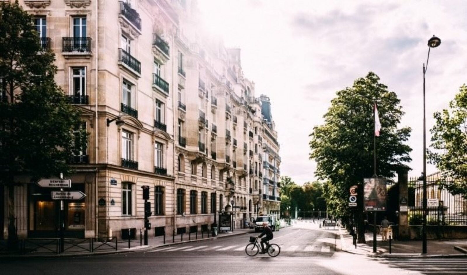 Un cycliste circule dans une rue calme de la ville bordée de bâtiments historiques et d'arbres, qui rappelle le quartier de rêve d'un courtier en prêt immobilier. Le soleil brille de mille feux, apportant une douce lumière sur la scène. En arrière-plan, davantage d’arbres et de bâtiments éloignés capturent l’atmosphère urbaine tranquille.