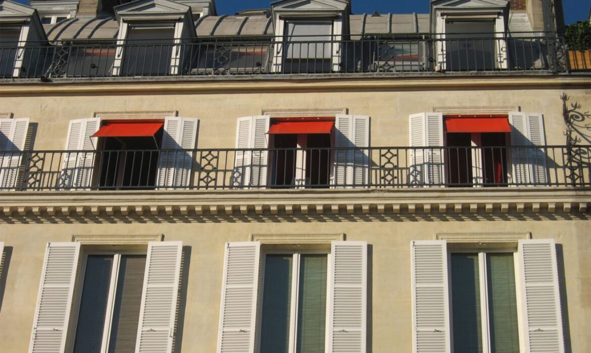 Un bâtiment beige avec des volets blancs, des auvents rouges et des balustrades en fer constitue un joyau de charme sur le marché immobilier. L'étage supérieur a des lucarnes avec un toit en pente ; les volets sont entièrement ouverts et les auvents rouges partiellement déployés, projetant des ombres sur le mur.