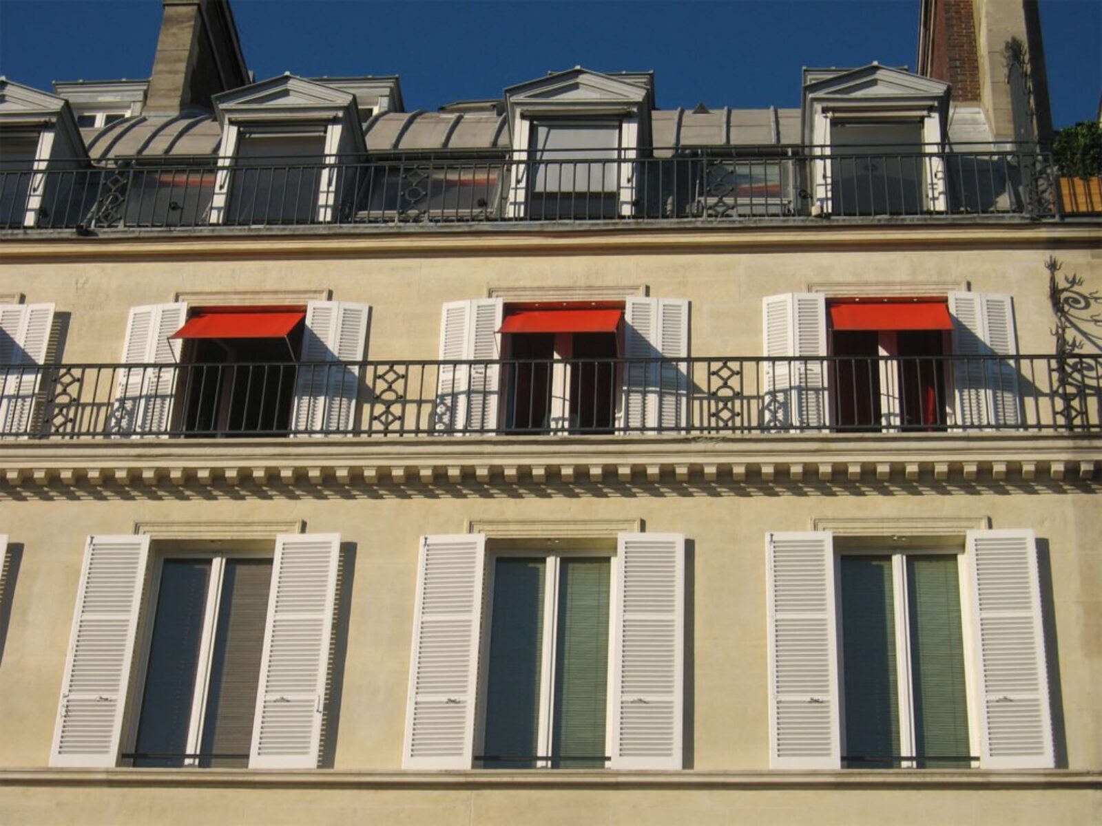 Un bâtiment beige avec des volets blancs, des auvents rouges et des balustrades en fer constitue un joyau de charme sur le marché immobilier. L'étage supérieur a des lucarnes avec un toit en pente ; les volets sont entièrement ouverts et les auvents rouges partiellement déployés, projetant des ombres sur le mur.