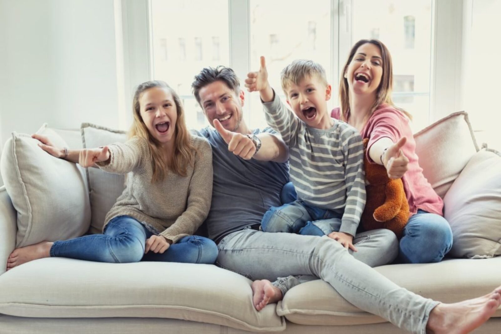 Une famille de quatre personnes est assise sur un canapé, souriante et montrant les pouces vers le haut. Les parents sont de chaque côté, avec deux enfants entre eux. La fille est à gauche, le garçon au milieu tient un ours en peluche. Ils apparaissent joyeux et détendus dans leur chambre lumineuse et luxueuse, reflétant leur goût pour l'immobilier de luxe.