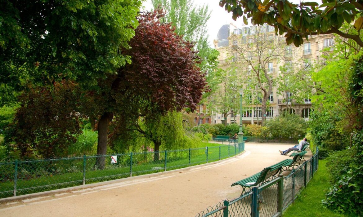 Une scène de parc sereine avec un chemin de gravier sinueux, une verdure luxuriante et un mélange d'arbres feuillus. Quelques bancs en bois bordent le chemin et quelques personnes peuvent être vues en train de se détendre dessus. Un bâtiment historique à l’architecture richement ornée est visible en arrière-plan.