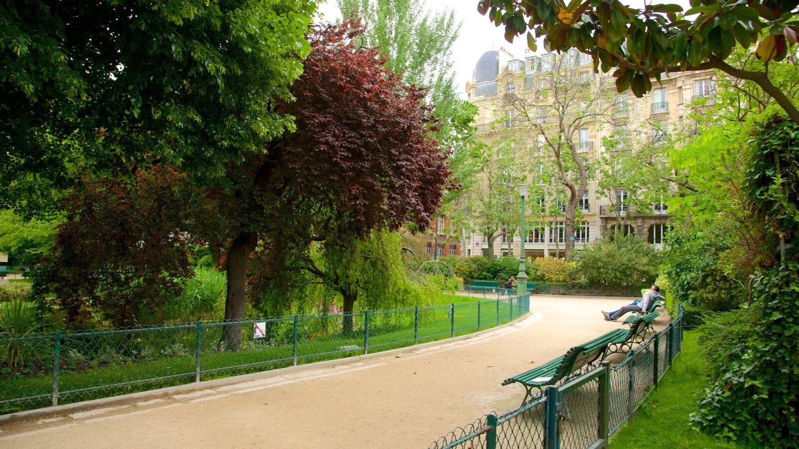 Une scène de parc sereine avec un chemin de gravier sinueux, une verdure luxuriante et un mélange d'arbres feuillus. Quelques bancs en bois bordent le chemin et quelques personnes peuvent être vues en train de se détendre dessus. Un bâtiment historique à l’architecture richement ornée est visible en arrière-plan.