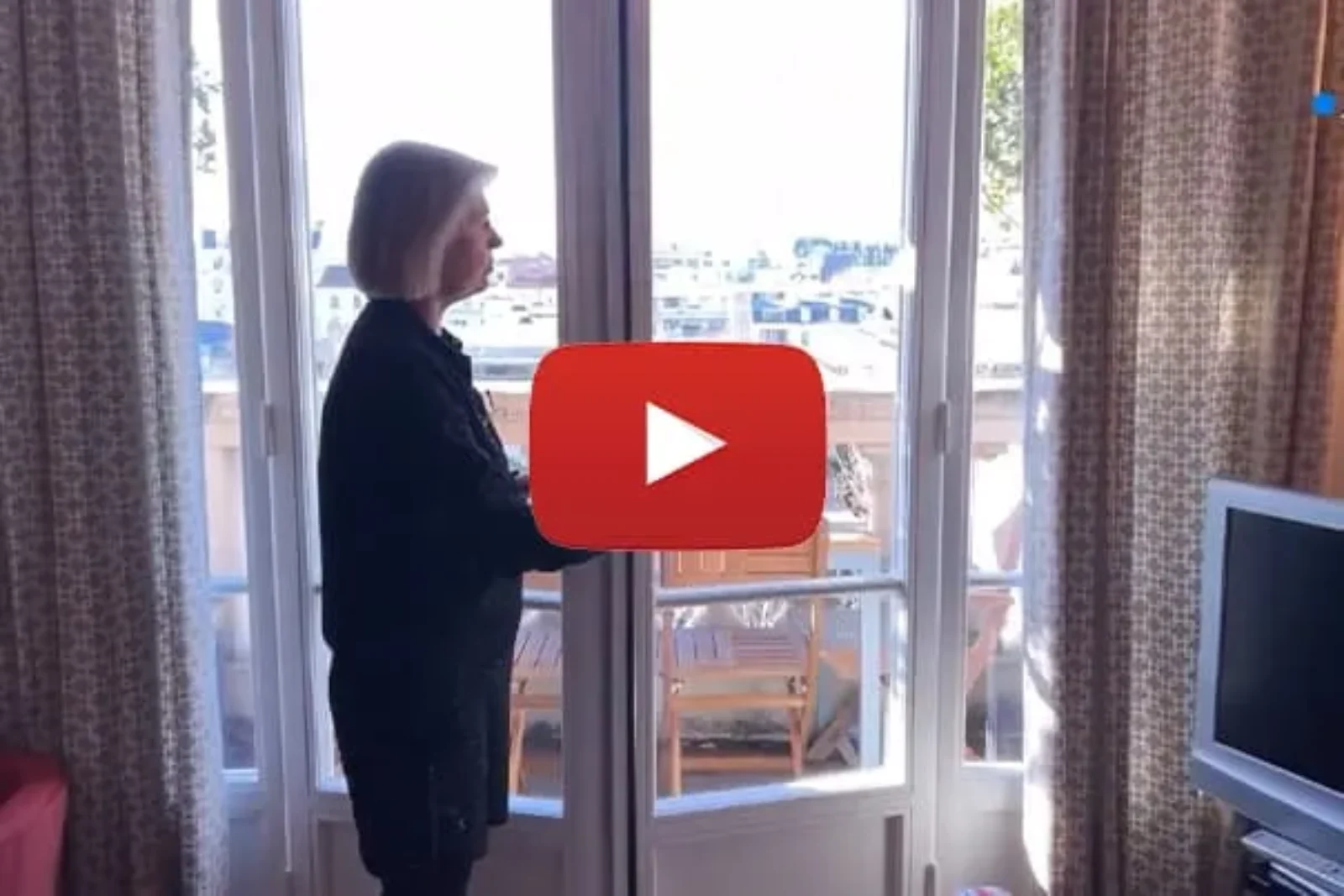 Une femme se tient face à de grandes portes vitrées et donne sur un balcon ensoleillé. La pièce intérieure comprend une télévision sur le côté droit de l’image. Un bouton de lecture rouge est superposé au centre, suggérant que l'image provient d'une vidéo.