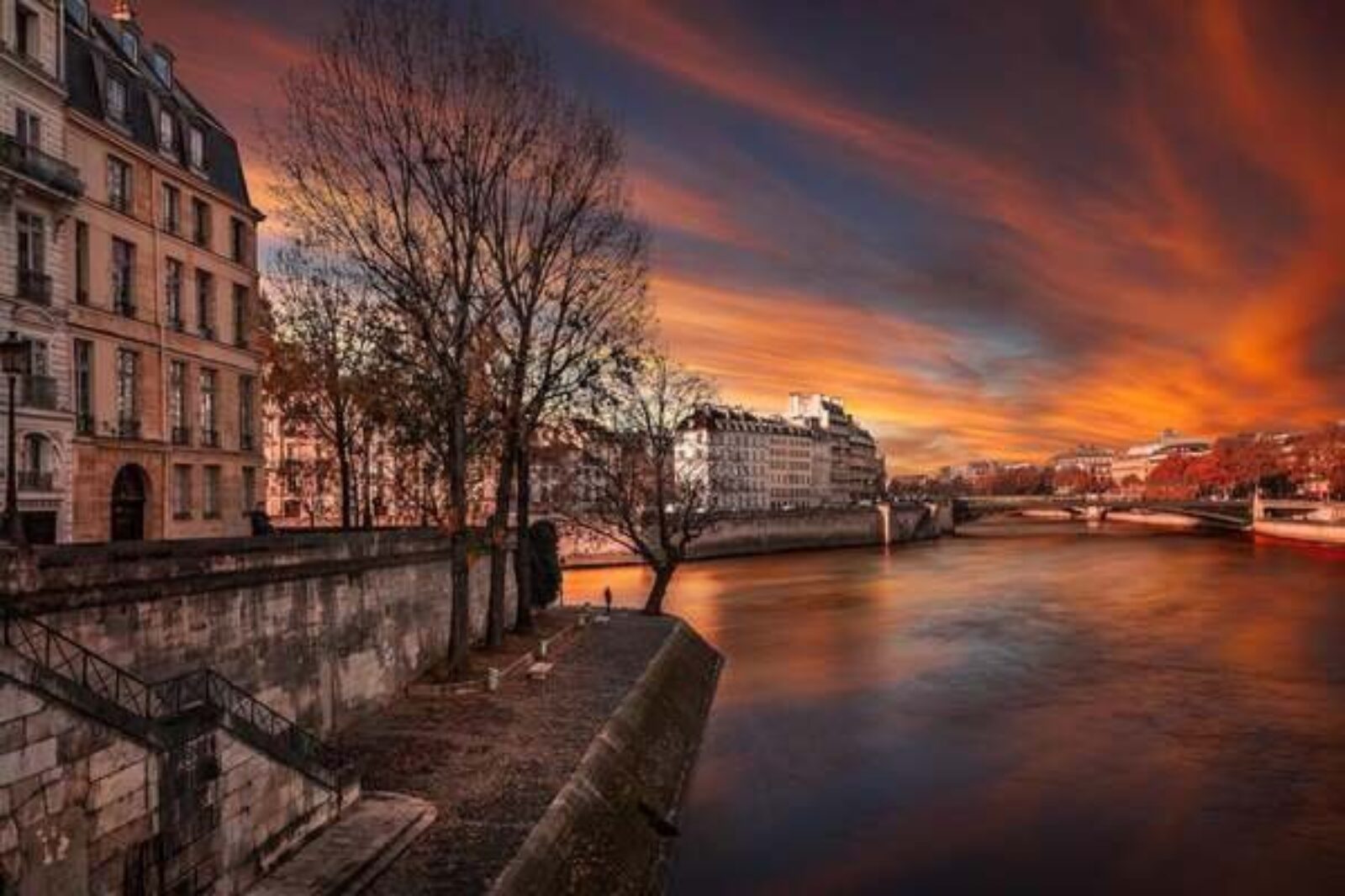 Une vue pittoresque sur une rivière dans une ville au coucher du soleil. Le ciel est rempli de teintes vibrantes d'orange, de rouge et de violet. Des bâtiments élégants bordent la rivière, des arbres aux feuilles d'automne et une rivière calme reflétant les couleurs du coucher du soleil. Un pont relie les deux rives, un décor idyllique pour tout Chasseur immobilier Ile-de-France.