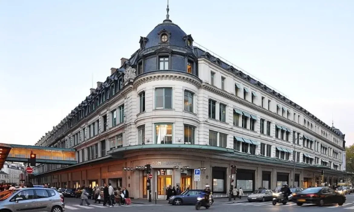 Un coin de rue animé avec un grand bâtiment historique à plusieurs étages doté d'une tour d'angle arrondie distinctive et de nombreuses fenêtres. Les piétons, les voitures et les scooters sont visibles, mettant en valeur une scène urbaine animée sur fond de grande architecture que vous pourriez trouver en tant que Chasseur d'appartement à Paris 7.