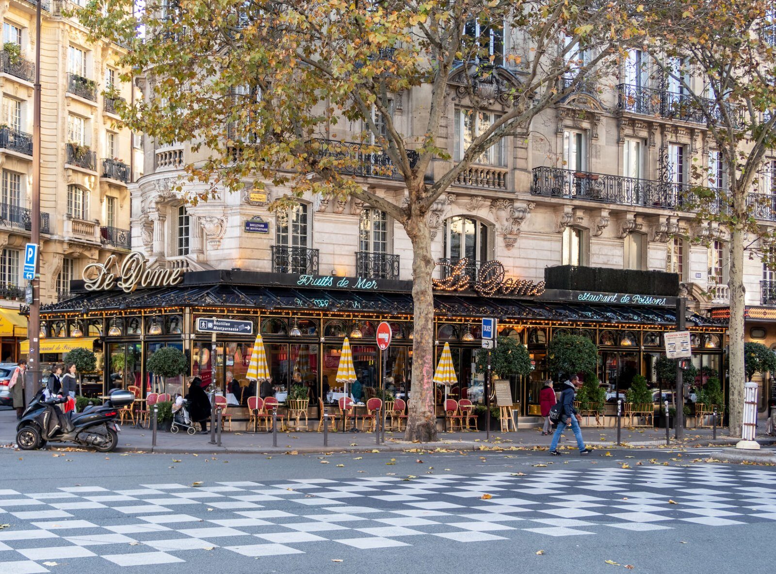 Vue du trottoir du Dôme Café à Paris, France. Le coin salon extérieur du café comprend des tables avec des chaises rouges et des parasols. Les gens passent et des arbres aux feuilles d’automne bordent la rue. Le bâtiment présente une architecture classique avec des balcons et des détails décoratifs, typiques des Chasseur Appartements Paris 14.