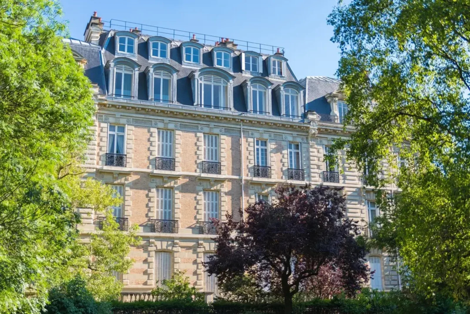 Un bâtiment français classique de plusieurs étages avec de hautes fenêtres cintrées, des balcons en fer forgé et un toit mansardé. La façade est de couleur claire avec des détails décoratifs en pierre. Des arbres au feuillage luxuriant vert et rouge foncé entourent le bâtiment, l’obscurcissant partiellement.