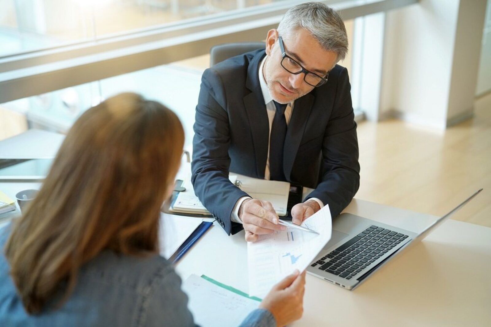 Un homme en costume d'affaires et lunettes est assis à un bureau en face d'une femme et lui montre un document relatif au plafonnement des loyers. Ils se trouvent dans un bureau bien éclairé avec de grandes fenêtres. La femme tient le document, tandis qu'un ordinateur portable et un presse-papiers sont visibles sur le bureau.